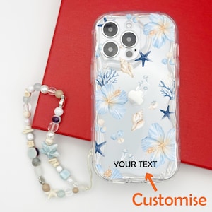 K&ouml;nnte beinhalten: Transparente Handyh&uuml;lle mit einem Blumen- und Muschelmuster. Die H&uuml;lle zeigt blaue Hibiskusbl&uuml;ten, Seesterne und Korallen. Der Text "YOUR TEXT" ist auf der H&uuml;lle aufgedruckt, mit einem Pfeil, der auf das Wort "Customise" zeigt. Eine Perlen-Handy-Schlaufe liegt daneben.