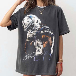 Könnte beinhalten: Dunkelgraues T-Shirt mit einem Footballspieler in einem Helm mit Sternlogo. Der Spieler trägt ein Trikot mit der Nummer 9. Das Shirt hat einen Used-Look und eine Unterschrift.