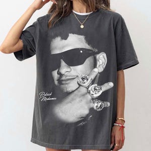 Op de afbeelding: Donkergrijs T-shirt met een zwart-wit afbeelding van een persoon met een zonnebril en ringen. De naam "Patrick Mahomes" is cursief geschreven. Het shirt heeft korte mouwen.