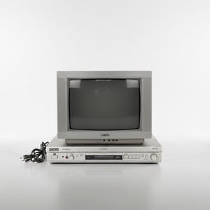 Puede incluir: Un televisor gris vintage se encuentra encima de un reproductor de DVD plateado. El televisor tiene una pantalla oscura y un diseño cuadrado. El reproductor de DVD tiene varios botones y una pantalla. Un cable de alimentación negro está conectado al reproductor de DVD.