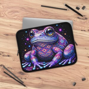 May include: A laptop sleeve with a colourful frog design. The frog is predominantly purple, with blue and pink details and intricate patterns. The background is a starry night. The sleeve has a zip closure. It is on a wooden surface.