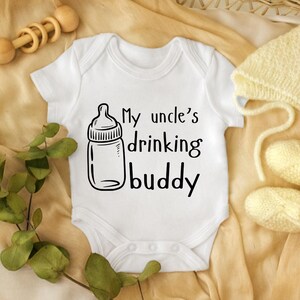 Puede incluir: Un body blanco para bebé con el texto "My uncle's drinking buddy" y una ilustración de biberón. El body está en una cesta de mimbre con un sonajero de madera, un gorro de punto y patucos. El fondo presenta una tela beige y vegetación.