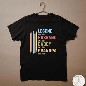 May include: Black t-shirt with the words "LEGEND, HUSBAND, DADDY, GRANDPA" in blue, red, yellow, and green. The shirt includes the years 1969, 1993, 1994, and 2025. The shirt is hanging on a metal hanger.
