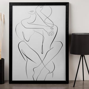 Pu&ograve; includere: Una stampa con cornice nera con un disegno a tratto minimalista di due figure che si abbracciano. L'opera d'arte &egrave; in nero su sfondo bianco. Sono visibili anche una lampada da tavolo nera con base a treppiede e un vaso nero con steli secchi.