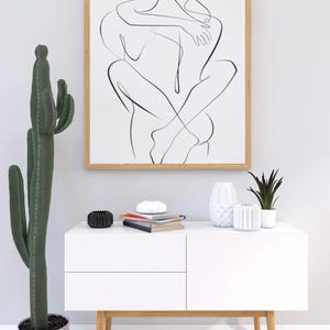 Pu&ograve; includere: Un'opera d'arte in bianco e nero incorniciata, raffigurante due figure che si abbracciano. L'opera &egrave; appesa sopra un mobile bianco con gambe in legno, adornato con vasi decorativi, libri e una piccola pianta in vaso. Un alto cactus a sinistra.