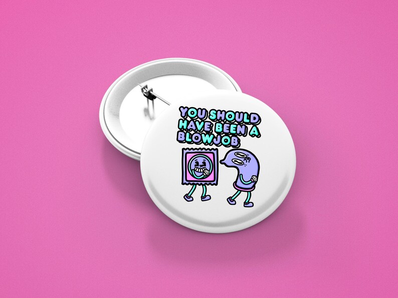 May include: White button badge with a pin back, featuring a cartoon design of a condom and a sperm cell with the text "YOU SHOULD HAVE BEEN A BLOW JOB" in blue and green. The badge is set against a pink background.