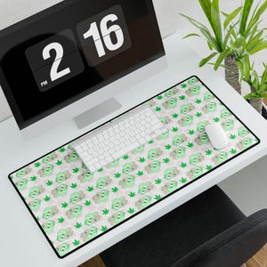 Puede incluir: Una alfombrilla de escritorio rectangular con borde negro presenta un patrón repetido de cerebros de dibujos animados verdes y hojas de cannabis sobre un fondo rosa claro. Un teclado y un ratón blancos están sobre la alfombrilla, con un monitor de ordenador al fondo.