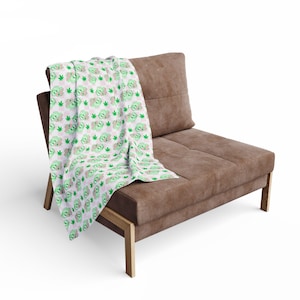May include: A white throw blanket with a repeating pattern of green cartoon characters, green cannabis leaves, and pink shapes. The blanket is draped over a brown sofa with wooden legs.