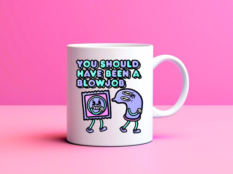May include: White ceramic mug with the text "YOU SHOULD HAVE BEEN A BLOWJOB" in teal and purple. The mug features cartoon illustrations of a condom and a sperm cell with legs, set against a pink background.