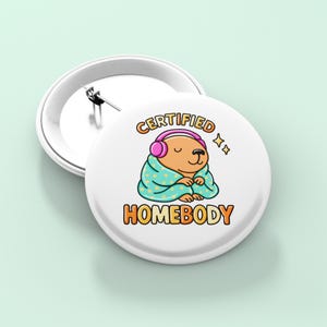 K&ouml;nnte beinhalten: Wei&szlig;er Ansteckbutton mit einem Cartoon-B&auml;ren, der rosa Kopfh&ouml;rer tr&auml;gt und in eine t&uuml;rkisfarbene Decke geh&uuml;llt ist. Der Text "CERTIFIED HOMEBODY" ist in Orange und Gelb dargestellt. Ein zweiter Button ist sichtbar.