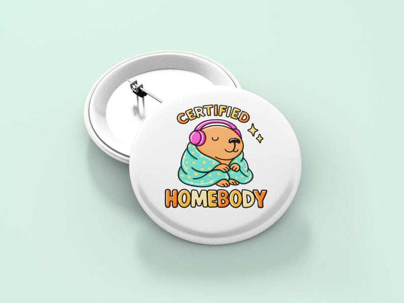 K&ouml;nnte beinhalten: Wei&szlig;er Ansteckbutton mit einem Cartoon-B&auml;ren, der rosa Kopfh&ouml;rer tr&auml;gt und in eine t&uuml;rkisfarbene Decke geh&uuml;llt ist. Der Text "CERTIFIED HOMEBODY" ist in Orange und Gelb dargestellt. Ein zweiter Button ist sichtbar.