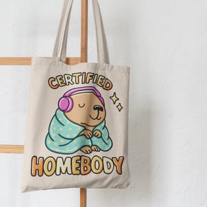 K&ouml;nnte beinhalten: Beige Tragetasche mit einem Cartoon-Wasserschwein, das Kopfh&ouml;rer tr&auml;gt und in eine Decke geh&uuml;llt ist. Der Text "CERTIFIED HOMEBODY" ist in bunten Buchstaben gedruckt. Das Design umfasst Sterne und ein gem&uuml;tliches Thema.
