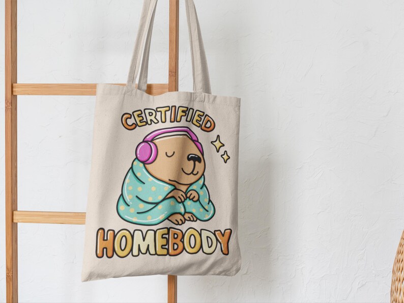 K&ouml;nnte beinhalten: Beige Tragetasche mit einem Cartoon-Wasserschwein, das Kopfh&ouml;rer tr&auml;gt und in eine Decke geh&uuml;llt ist. Der Text "CERTIFIED HOMEBODY" ist in bunten Buchstaben gedruckt. Das Design umfasst Sterne und ein gem&uuml;tliches Thema.