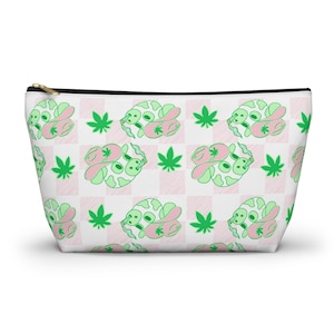 May include: White cosmetic bag featuring a repeating pattern of green and pink cartoon characters, cannabis leaves, and a pink and white checkered background. The bag has a black zip and trim.
