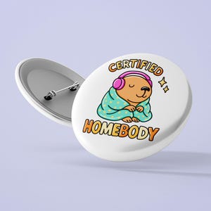 K&ouml;nnte beinhalten: Wei&szlig;er Button mit einem Cartoon-B&auml;ren, der rosa Kopfh&ouml;rer und eine t&uuml;rkisfarbene Decke tr&auml;gt. Der Text auf dem Button lautet "CERTIFIED HOMEBODY" in Orange und Gelb. Ein zweiter Button ist sichtbar.