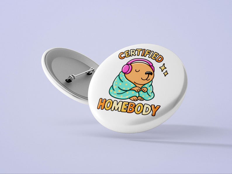 K&ouml;nnte beinhalten: Wei&szlig;er Button mit einem Cartoon-B&auml;ren, der rosa Kopfh&ouml;rer und eine t&uuml;rkisfarbene Decke tr&auml;gt. Der Text auf dem Button lautet "CERTIFIED HOMEBODY" in Orange und Gelb. Ein zweiter Button ist sichtbar.