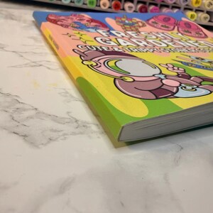 Puede incluir: Un libro colorido titulado "Fresh Gardens: Cute & Trippy Stickers" con ilustraciones de personajes de dibujos animados. El libro tiene un lomo amarillo y una portada rosa claro con detalles en azul y verde. El libro est&aacute; sobre una superficie de m&aacute;rmol blanco.