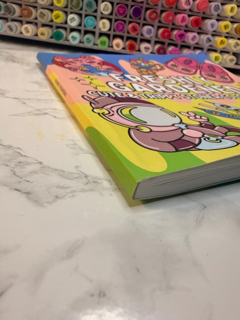 Puede incluir: Un libro colorido titulado "Fresh Gardens: Cute & Trippy Stickers" con ilustraciones de personajes de dibujos animados. El libro tiene un lomo amarillo y una portada rosa claro con detalles en azul y verde. El libro est&aacute; sobre una superficie de m&aacute;rmol blanco.