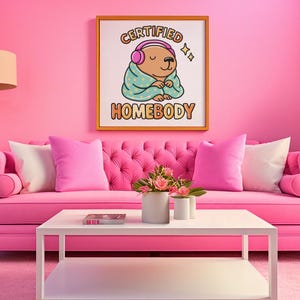 K&ouml;nnte beinhalten: Ein rosa getuftetes Sofa mit passenden Kissen und einem wei&szlig;en Couchtisch. Ein gerahmter Druck an der Wand zeigt "CERTIFIED HOMEBODY" mit einem Cartoon-B&auml;ren mit Kopfh&ouml;rern. Der Raum ist in Rosat&ouml;nen gehalten.