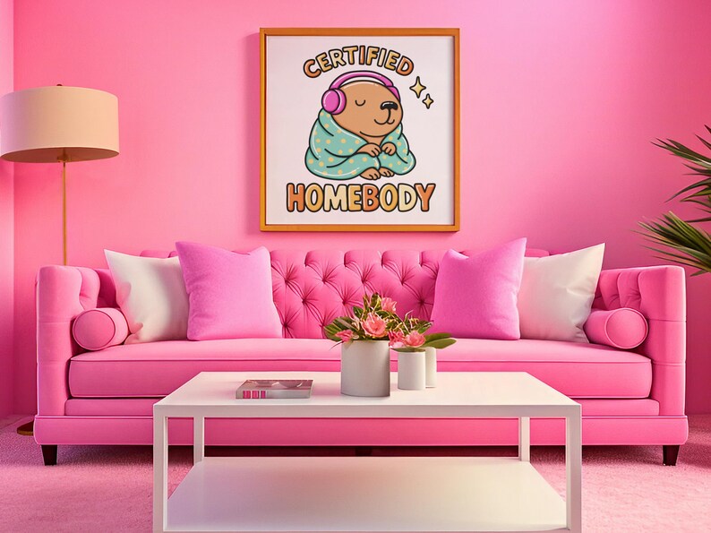 K&ouml;nnte beinhalten: Ein rosa getuftetes Sofa mit passenden Kissen und einem wei&szlig;en Couchtisch. Ein gerahmter Druck an der Wand zeigt "CERTIFIED HOMEBODY" mit einem Cartoon-B&auml;ren mit Kopfh&ouml;rern. Der Raum ist in Rosat&ouml;nen gehalten.