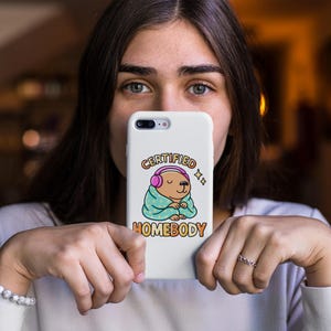 K&ouml;nnte beinhalten: Wei&szlig;es Handycase mit einem Cartoon-B&auml;ren, der rosa Kopfh&ouml;rer und eine t&uuml;rkisfarbene Decke tr&auml;gt. Der Text auf dem Case lautet "CERTIFIED HOMEBODY" in einer bunten Schriftart. Das Case wird von einer Person gehalten.