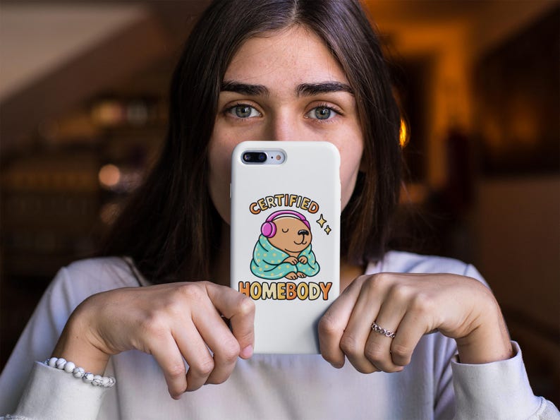 K&ouml;nnte beinhalten: Wei&szlig;es Handycase mit einem Cartoon-B&auml;ren, der rosa Kopfh&ouml;rer und eine t&uuml;rkisfarbene Decke tr&auml;gt. Der Text auf dem Case lautet "CERTIFIED HOMEBODY" in einer bunten Schriftart. Das Case wird von einer Person gehalten.
