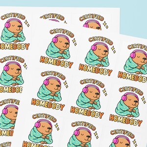 K&ouml;nnte beinhalten: Bl&auml;tter mit Aufklebern mit einem Cartoon-B&auml;ren mit rosa Kopfh&ouml;rern und in eine t&uuml;rkisfarbene Decke geh&uuml;llt. Die Aufkleber tragen den Schriftzug "CERTIFIED HOMEBODY" in orange und wei&szlig;, mit Sternakzenten.