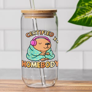 K&ouml;nnte beinhalten: Klarer Glasbecher mit Bambusdeckel und Strohhalm. Der Becher zeigt eine Cartoon-Illustration eines braunen B&auml;ren mit rosa Kopfh&ouml;rern und einer t&uuml;rkisfarbenen Decke mit wei&szlig;en Punkten. Der Text auf dem Becher lautet "CERTIFIED HOMEBODY".