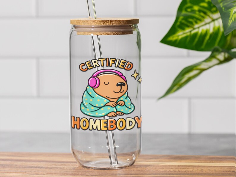 K&ouml;nnte beinhalten: Klarer Glasbecher mit Bambusdeckel und Strohhalm. Der Becher zeigt eine Cartoon-Illustration eines braunen B&auml;ren mit rosa Kopfh&ouml;rern und einer t&uuml;rkisfarbenen Decke mit wei&szlig;en Punkten. Der Text auf dem Becher lautet "CERTIFIED HOMEBODY".