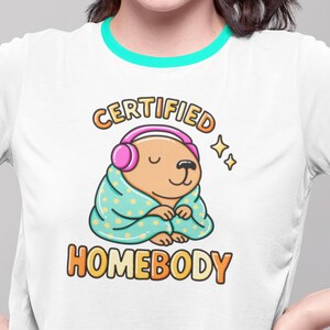 K&ouml;nnte beinhalten: Wei&szlig;es T-Shirt mit t&uuml;rkisfarbenem Besatz und einer Grafik eines Cartoon-Wasserschweins mit Kopfh&ouml;rern und in eine Decke geh&uuml;llt. Der Text lautet "CERTIFIED HOMEBODY" in Orange und Gelb.
