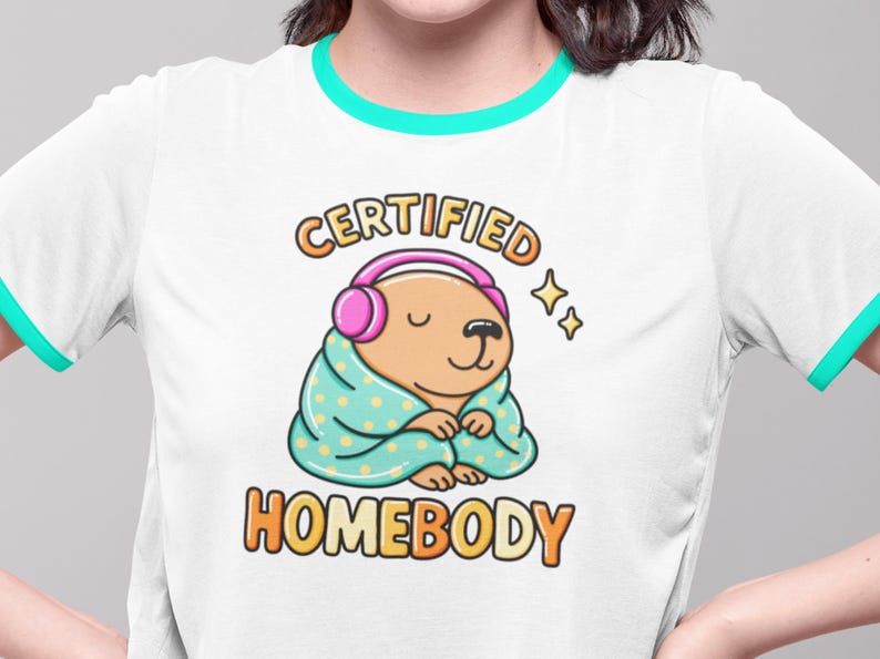 K&ouml;nnte beinhalten: Wei&szlig;es T-Shirt mit t&uuml;rkisfarbenem Besatz und einer Grafik eines Cartoon-Wasserschweins mit Kopfh&ouml;rern und in eine Decke geh&uuml;llt. Der Text lautet "CERTIFIED HOMEBODY" in Orange und Gelb.