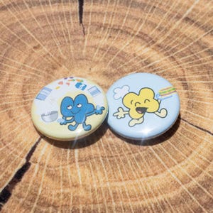 May include: Two round pinback buttons with cartoon characters on a light yellow and light blue background. One button features a blue character, the other a yellow character. The buttons are on a wooden surface.