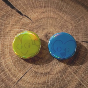 May include: Two circular, iridescent buttons featuring cartoon faces. One button is lime green, the other is blue. The buttons are on a wooden surface with a natural grain pattern. The cartoon faces have large eyes and smiling mouths.