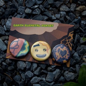 May include: A set of three circular pin-back buttons attached to a rectangular display card labeled EARTH ELEMETAL PIN SET. The pins feature a geometric orange and black pattern, a smiling yellow face with wavy hair, and a dark cracked earth design.