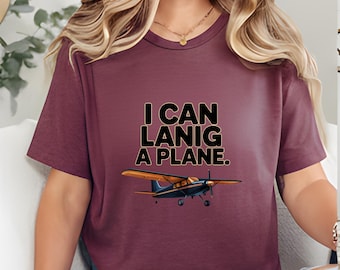 May include: Burgundy t-shirt with the text "I CAN LANIG A PLANE." and an illustration of a small airplane with blue and gold accents. The shirt is a crew neck style.