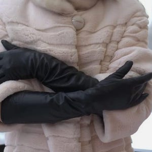 May include: A pair of long black leather gloves are worn by a person wearing a white fur coat. The gloves are folded over each other and the person's arms are crossed.