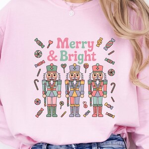 May include: Pink sweatshirt with the text "Merry & Bright" and a graphic of three nutcrackers. The design includes candy canes, lollipops, and other sweets. The nutcrackers are in pastel colors.