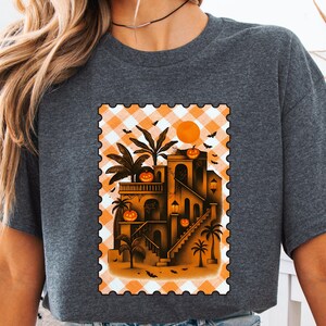 May include: Dark gray t-shirt featuring a Halloween-themed graphic. The graphic depicts a building with palm trees, pumpkins, and bats, set against an orange and white checkered background. The design is framed like a postage stamp.