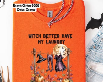 May include: Orange t-shirt with the text "Witch Better Have My Laundry." The shirt features a Halloween-themed graphic of witch clothing hanging on a clothesline, with pumpkins and a full moon. A silver chain necklace is draped over the shirt.