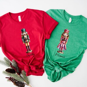 May include: Two short-sleeved t-shirts, one red and one green. The red shirt features a nutcracker illustration with the word "Daddy" below. The green shirt has a similar nutcracker design with "Mama" below.