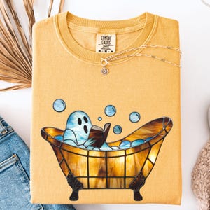 May include: Mustard yellow Comfort Colors sweatshirt featuring a cartoon ghost relaxing in a stained-glass bathtub and reading a book. The design includes blue bubbles. A gold necklace with a sun pendant is draped on the shirt.