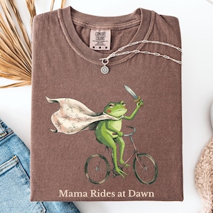 May include: Brown Comfort Colors t-shirt with a graphic of a green frog riding a bicycle and holding a knife. The frog wears a cape and the text "Mama Rides at Dawn" is below the graphic. A silver necklace is draped across the shirt.