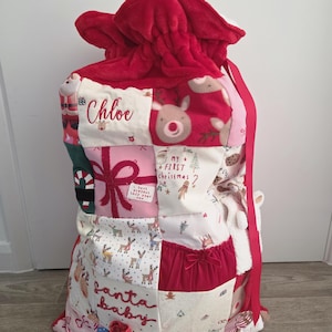 Könnte beinhalten: Ein Patchwork-Weihnachtsgeschenksack mit rotem Plüsch-Kordelzug und verschiedenen Stoffquadraten mit Weihnachtsmotiven wie Rentieren, Geschenken und Keksen. Der Text Chloe, My First Christmas und Santa baby ist auf den Stoff gedruckt.