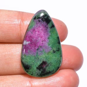 Può includere: Una gemma a forma di goccia con una superficie lucida. La pietra presenta un vivace mix di colori, tra cui un'area centrale di rosa brillante, circondata dal verde e accentuata da macchie nere. La gemma è tenuta in una mano.