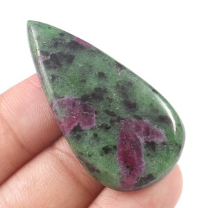 Könnte beinhalten: Ein polierter, tropfenförmiger Edelstein mit einer leuchtend grünen Basis und dunklen schwarzen und tiefroten Einschlüssen. Der Stein ist glatt und reflektierend und zeigt natürliche Muster und Farbvariationen. Der Edelstein wird in einer Hand gehalten.