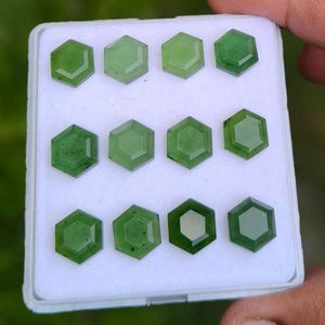 May include: A collection of twelve hexagonal green gemstones arranged in a grid pattern inside a white rectangular display box. Each stone features a faceted surface with a smooth, polished finish and varying shades of translucent forest green.