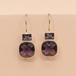 Könnte beinhalten: Ein Paar goldfarbene Ohrhänger. Jeder Ohrring hat einen kleinen, quadratischen, lila Edelstein über einem größeren, kissenförmig geschliffenen, lila Edelstein. Die Ohrringe hängen an einem einfachen goldenen Haken.