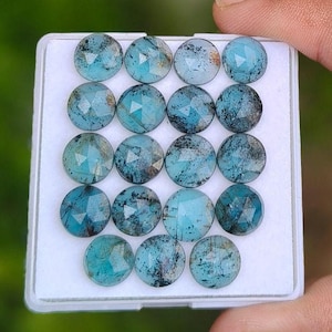 May include: A collection of seventeen round, faceted blue gemstones arranged in a grid on a white display tray. Each stone features a distinct teal-blue hue with dark, natural inclusions and a polished, multi-faceted surface reflecting light.