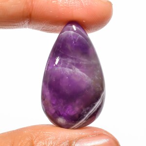 Könnte beinhalten: Ein polierter, tropfenförmiger Amethyst-Edelstein. Der Edelstein zeigt einen tiefvioletten Farbton mit helleren, durchscheinenden Streifen und Wirbeln. Der Stein wird zwischen zwei Fingern vor weißem Hintergrund gehalten.