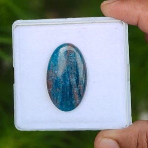 May include: An oval-shaped, polished gemstone featuring a vibrant blue color with visible reddish-brown inclusions and a smooth, reflective surface. The stone is centered on a square white display pad, showcasing its natural texture and color variations.